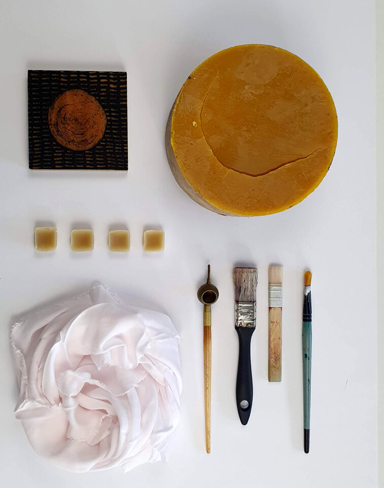 Na zdjęciu widoczne są przedmioty potrzebne do stworzenia batiku. Jest bryła wosku, biały materiał, pędzle i tjanting - narzędzie do malowania woskiem.